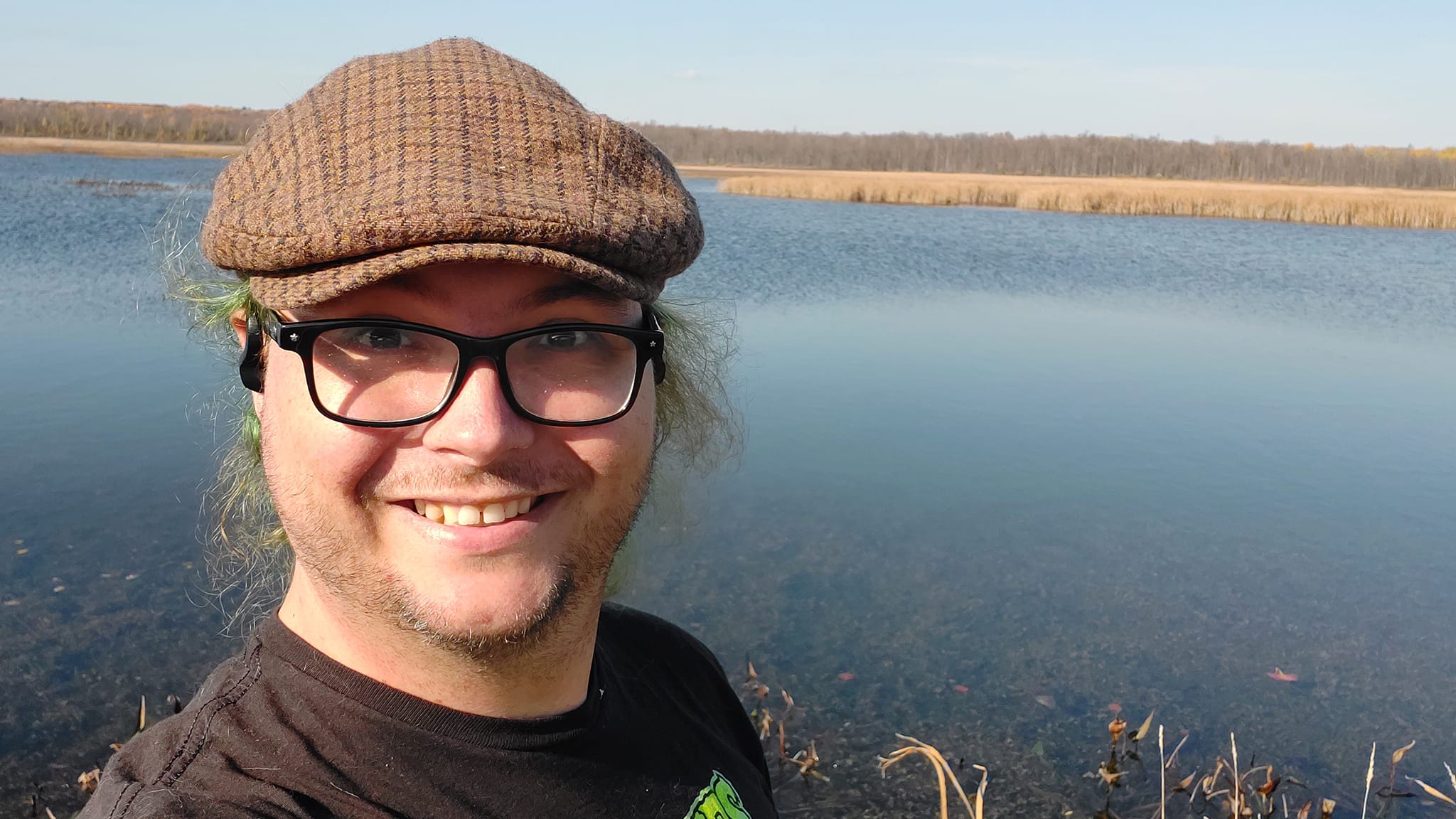 A man is wearing a brown flat cap and black shirt, standing in front of the open water of a marsh